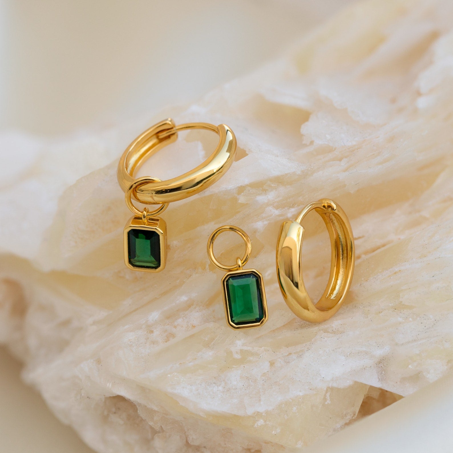 Dangling Emerald Birthstone Hoops rest on a light-colored textured surface.