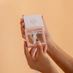 Hands hold a Birthstone Mystery Box with a gold necklace and earrings featuring green gemstones against a peach background—a personalized gift perfect for any occasion.