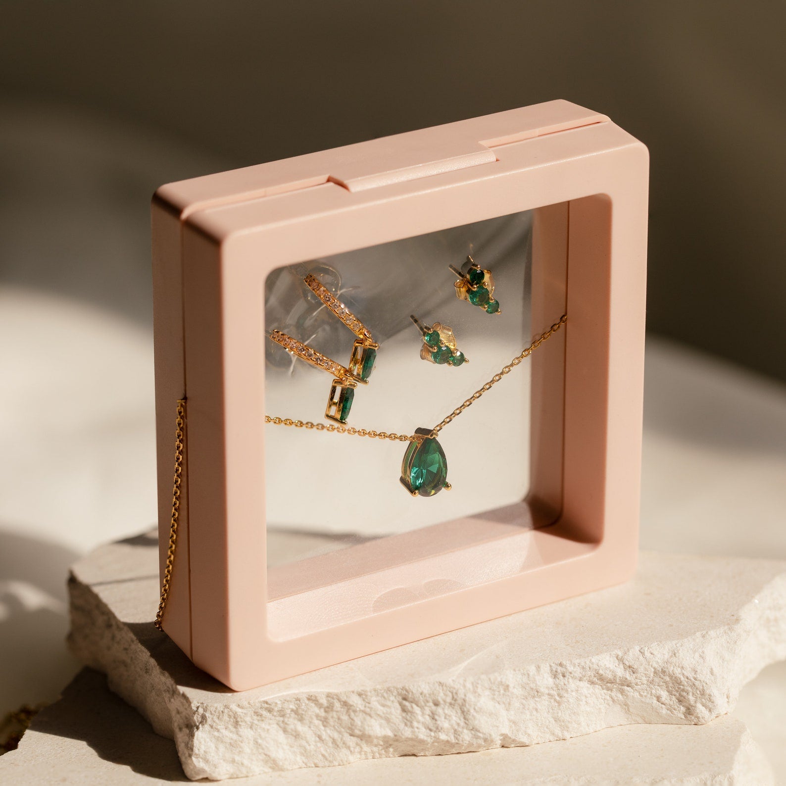 Birthstone Mystery Box featuring a gold and green gemstone necklace and earrings set, elegantly displayed in a pink frame atop white textured stones.