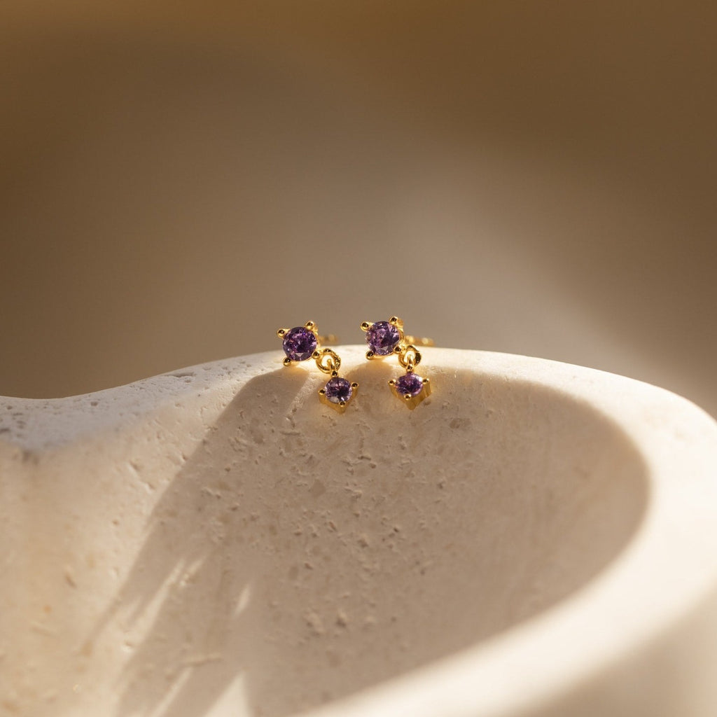 Amethyst Birthstone Drop Studs are showcased on a textured, light-colored stone surface, featuring petite purple gems.
