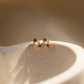 Amethyst Birthstone Drop Studs are showcased on a textured, light-colored stone surface, featuring petite purple gems.