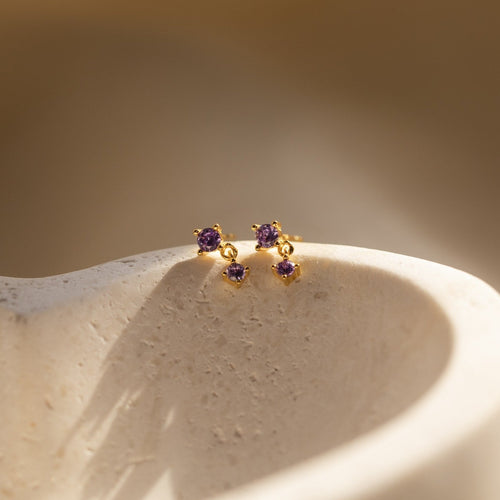 Amethyst Drop Studs with small purple gemstones rest on a textured, light-colored surface in soft sunlight.