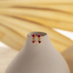 Ruby Birthstone Drop Studs featuring red gemstones displayed on a smooth white vase against a soft beige background.