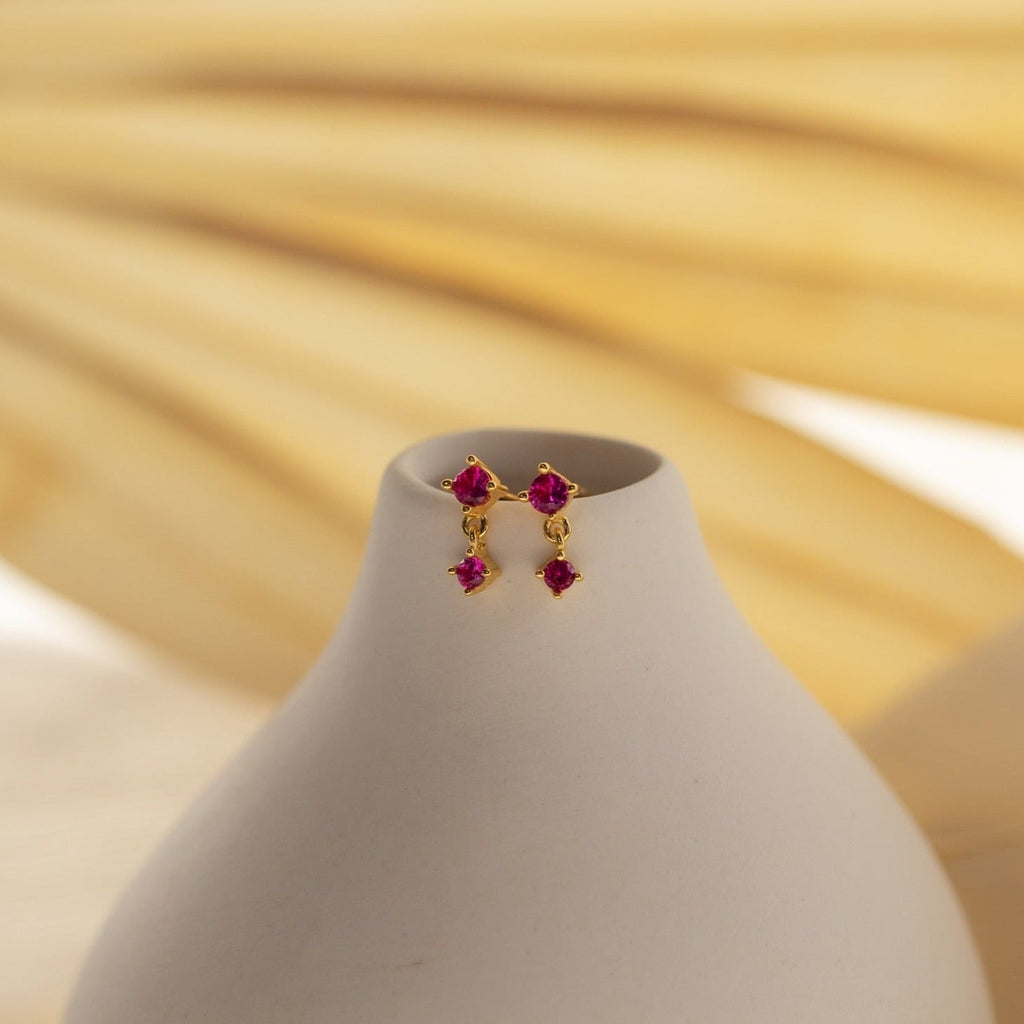 The Opal Birthstone Drop Studs, featuring dainty red gemstones, are displayed on a smooth white vase set against a soft beige background.