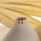 The Opal Birthstone Drop Studs, featuring dainty red gemstones, are displayed on a smooth white vase set against a soft beige background.
