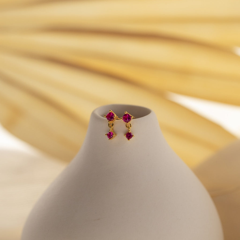 Emerald Birthstone Drop Studs—dainty gold earrings with three small green gemstones on a white stand, ideal as birthstone drop earrings, shown against a soft beige background.