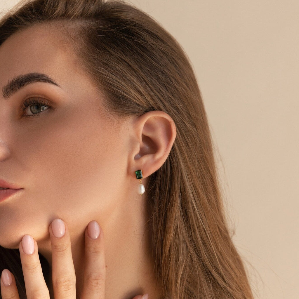 A woman with long brown hair gently touches her chin while wearing Emerald Pearl Drop Earrings.