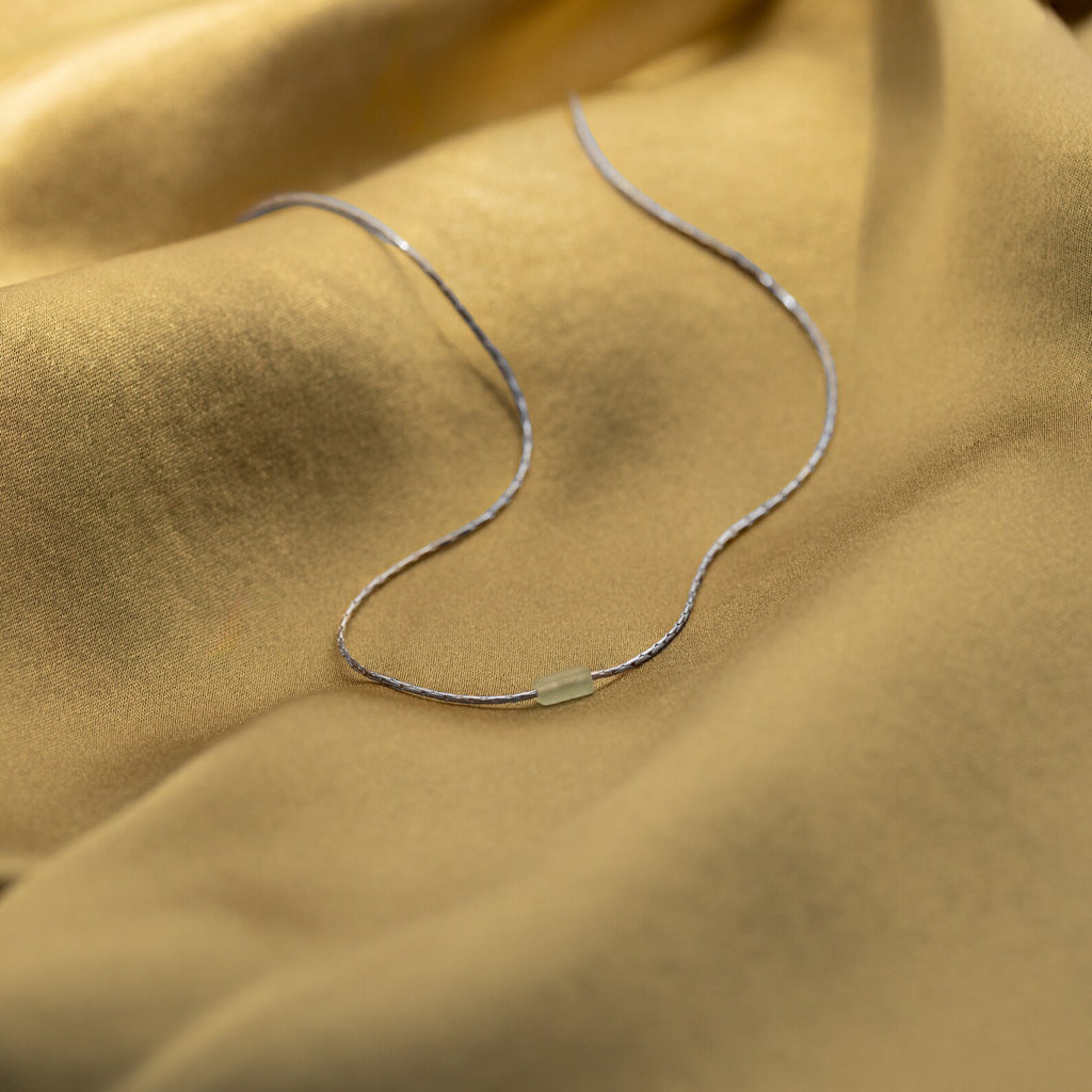 The Dainty Aventurine Bar Necklace, featuring a slim silver chain and small aventurine bar, rests gently on soft gold fabric.