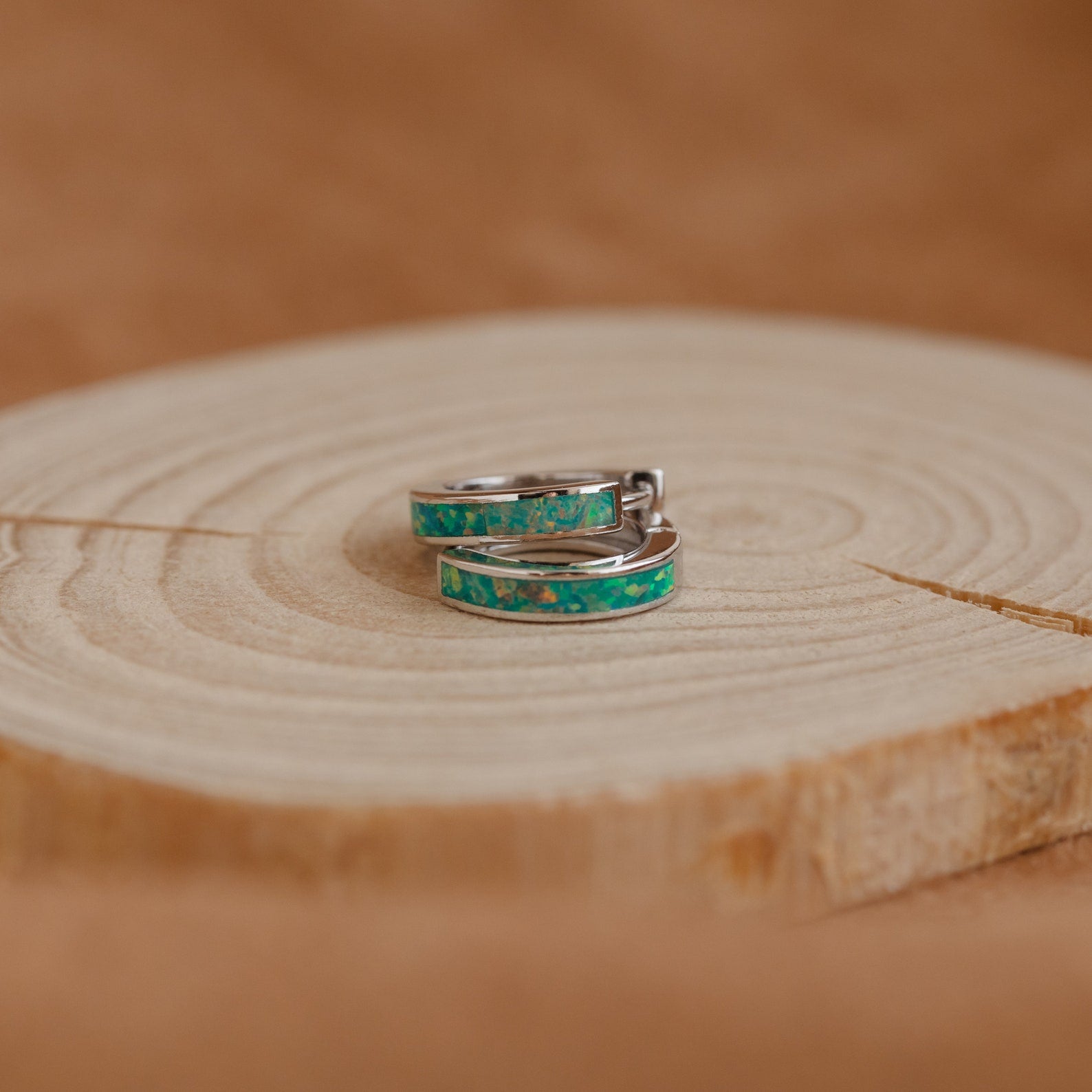 Green Opal Inlay Huggies, delicate silver hoop earrings with garden green opal inlay, are displayed on a round light wooden surface with a softly blurred brown background.
