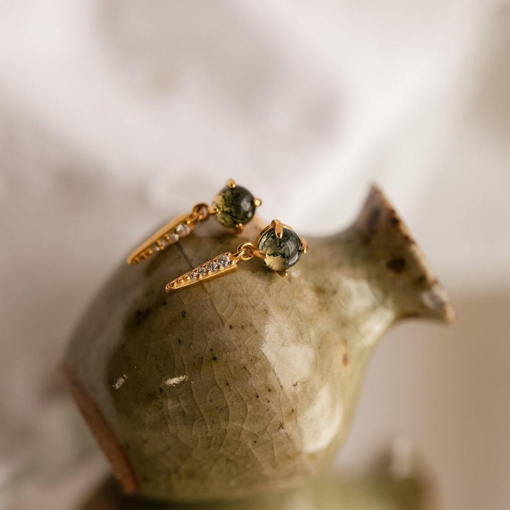 The Agate Diamond Drop Earrings feature dangling gold with green agate stones and sparkling diamonds, beautifully displayed on a glossy ceramic vase.