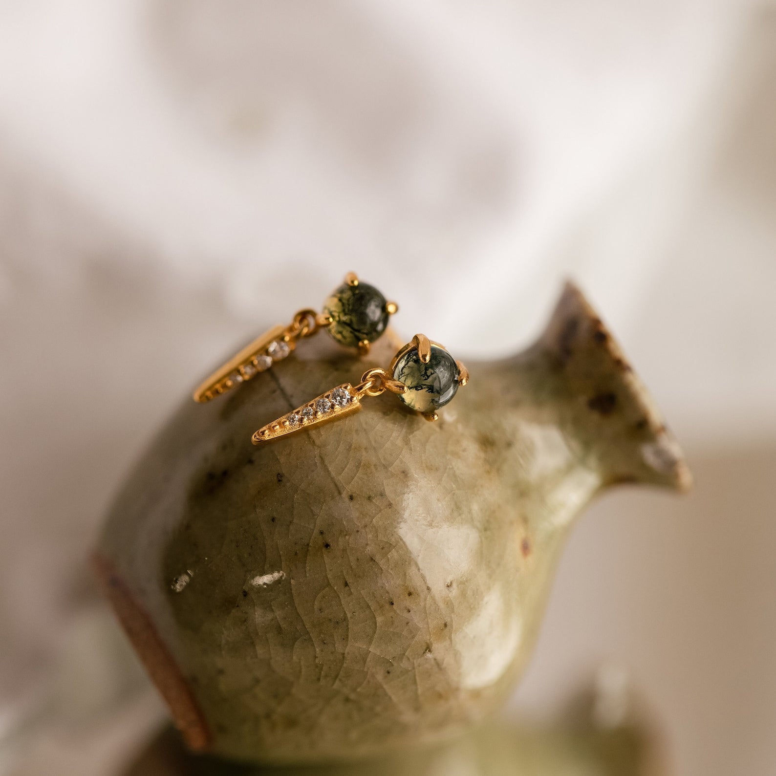 The Agate Diamond Drop Earrings feature dangling gold with green agate stones and sparkling diamonds, beautifully displayed on a glossy ceramic vase.