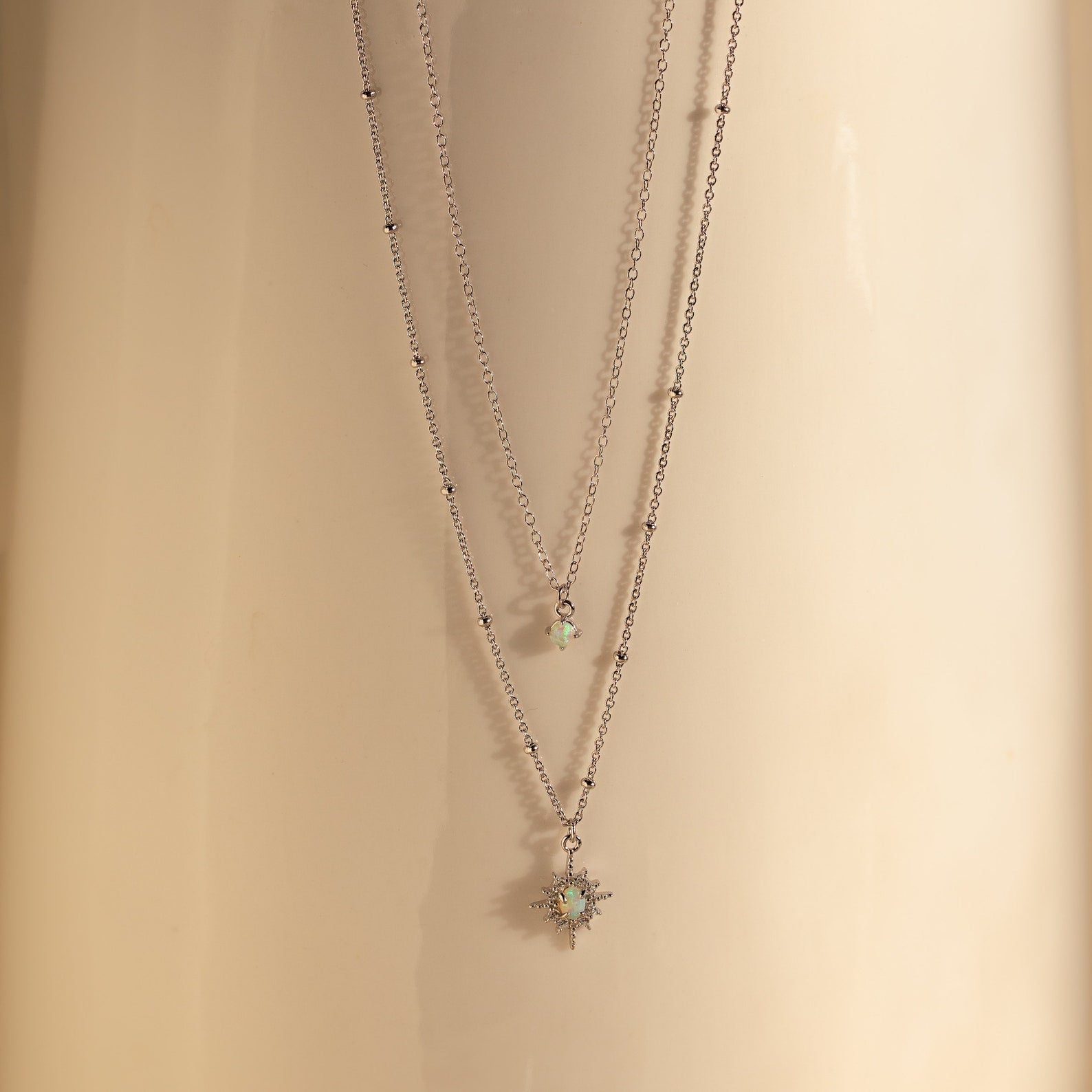 The Opal Star Duo Necklace features two delicate gold chains with opal pendants—one displaying a subtle starburst design—arranged against a smooth beige background for a stunning layered look.
