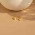 Dainty Opal Teardrop Studs—gold earrings featuring pearl teardrop centers and small crystals—are displayed on a beige surface, offering an elegant look.