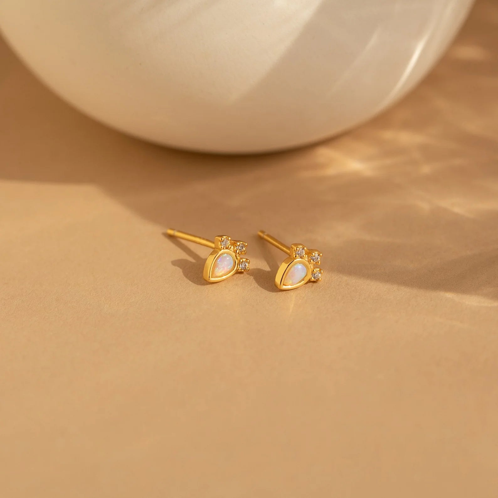 The Opal Drop Earrings Set features gold studs with pearl-like teardrop centers and gemstone accents, evoking elegance and perfect for layering jewelry on a beige surface.