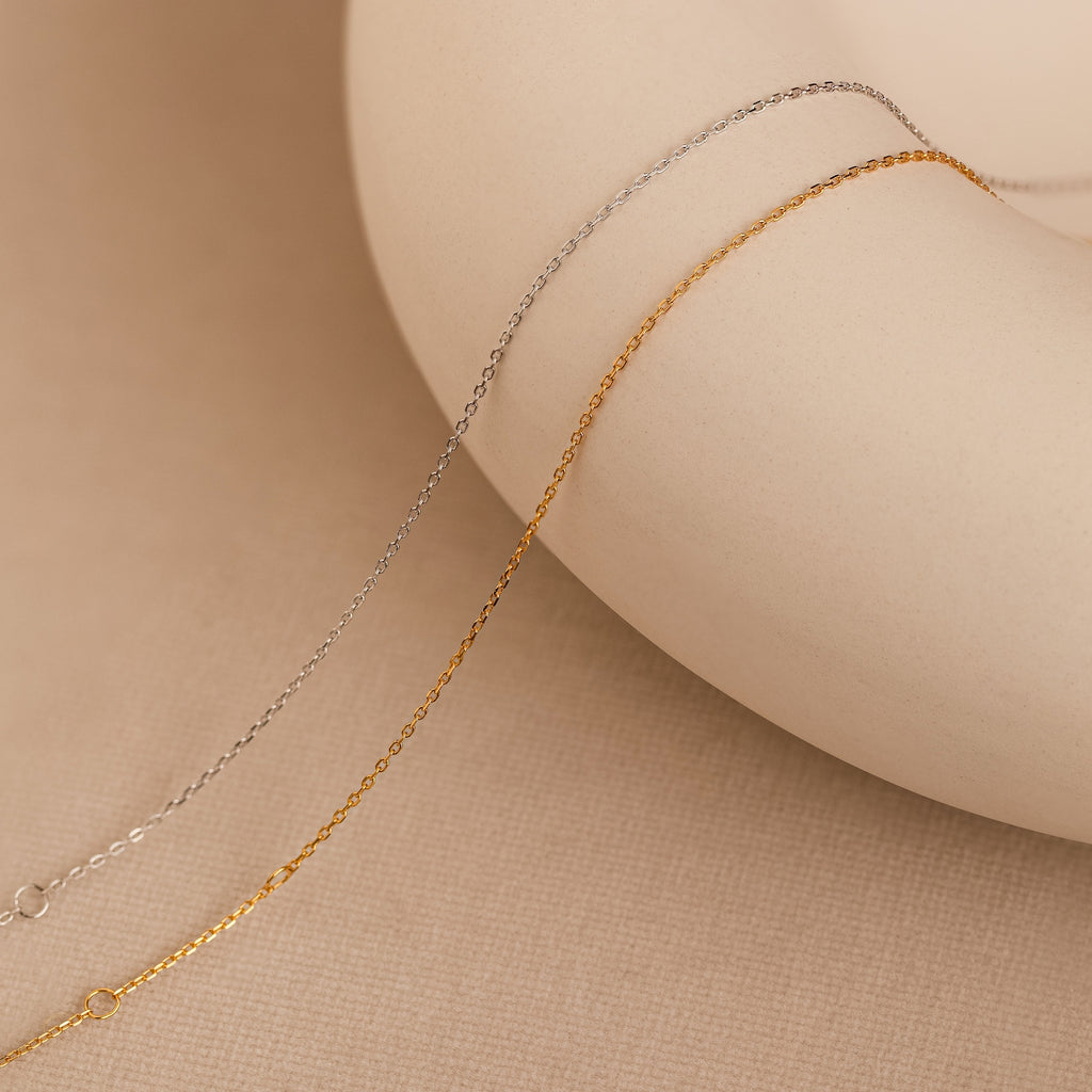 The Micro Rolo Chain Anklet by Caitlyn Minimalist in 18K gold and sterling silver drapes elegantly over a round beige object on a textured beige surface.