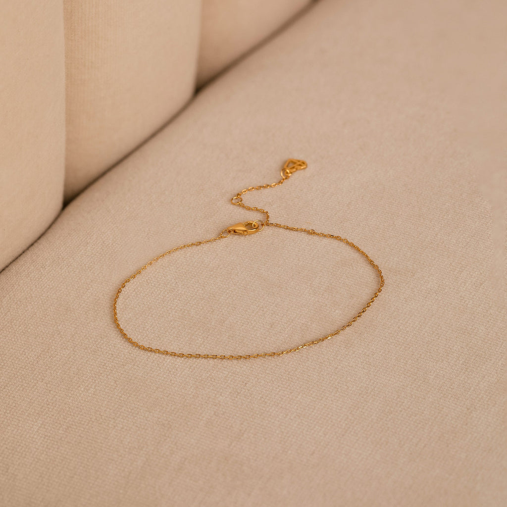 The Micro Rolo Chain Anklet by Caitlyn Minimalist, crafted in delicate 18K gold with a micro rolo chain and clasp, rests on a beige fabric surface.