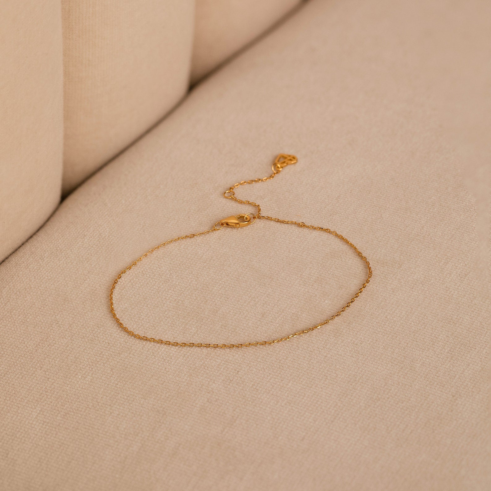 The Micro Rolo Chain Anklet by Caitlyn Minimalist, crafted in delicate 18K gold with a micro rolo chain and clasp, rests on a beige fabric surface.