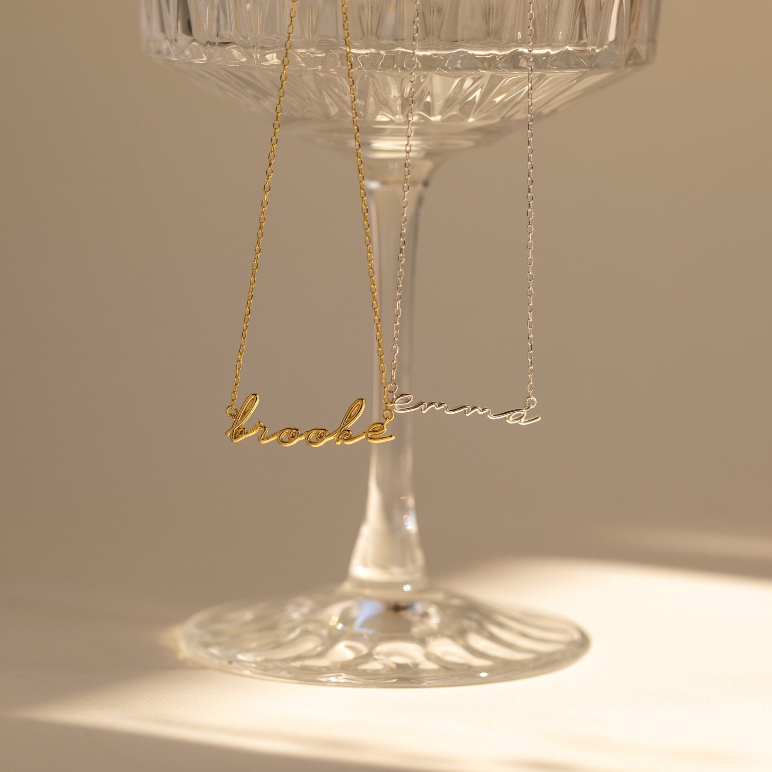 Two Dainty Bubble Name Necklaces—one gold reading “brooke” and one silver reading “emma”—hang on a clear glass cup.