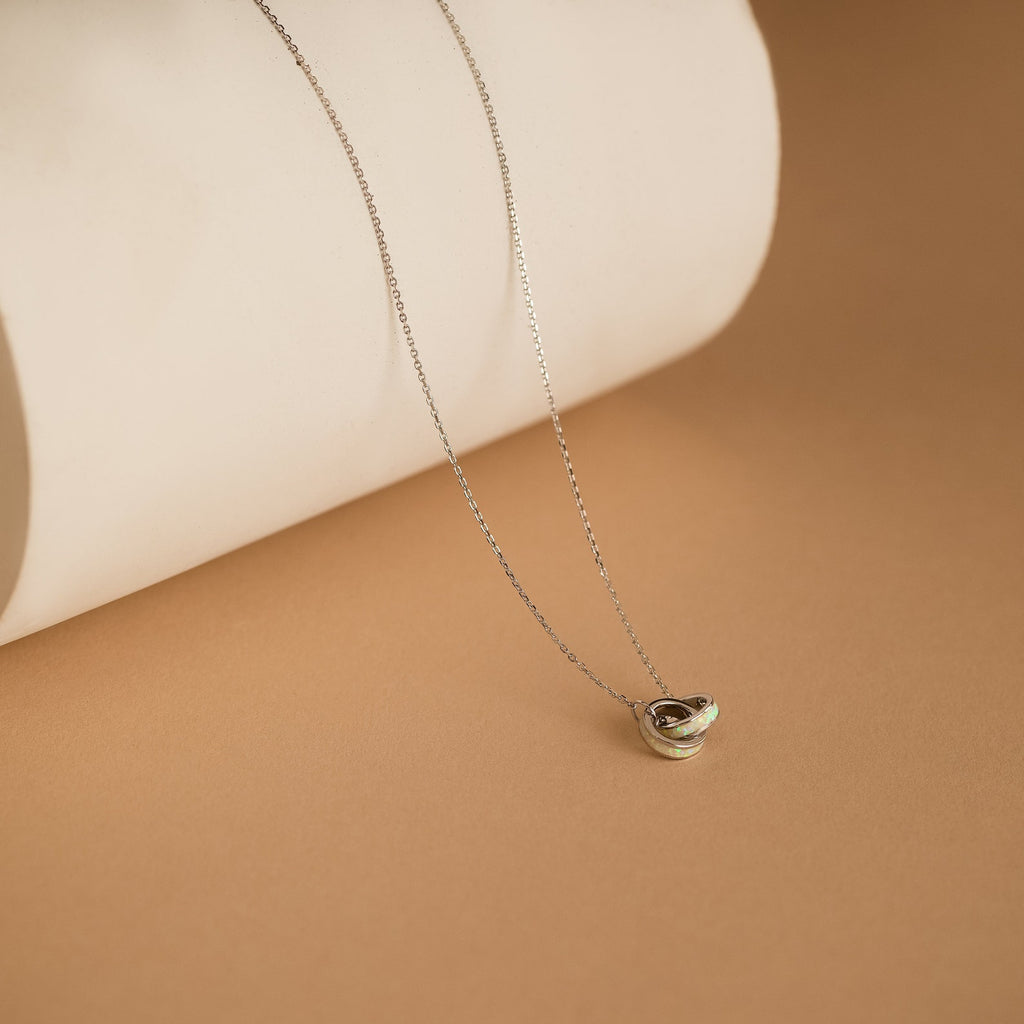 Interlocking Opal Inlay Necklace with a small pendant, showcased on a beige surface against a rolled white paper background—a stunning symbol of endless love, ideal for celebrating anniversaries.