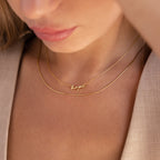 Close-up of a woman wearing gold necklaces, featuring the Tiny Venice Name Necklace personalized with her name, alongside another necklace displaying "hope" in cursive script.