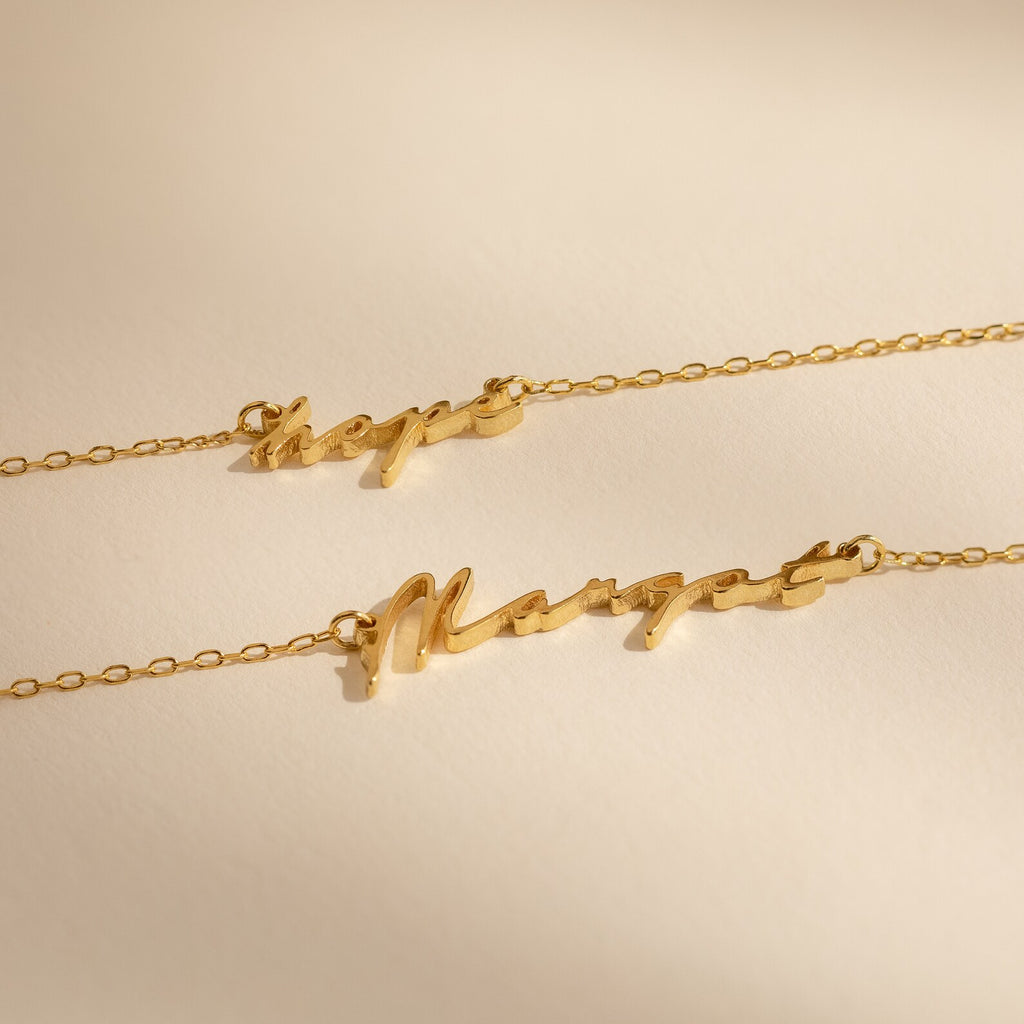 Two gold chain necklaces, one being the Tiny Venice Name Necklace with cursive nameplate charms reading "Hope" and "Margot," displayed on a neutral background.