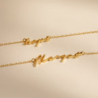 Two gold chain necklaces, one being the Tiny Venice Name Necklace with cursive nameplate charms reading "Hope" and "Margot," displayed on a neutral background.