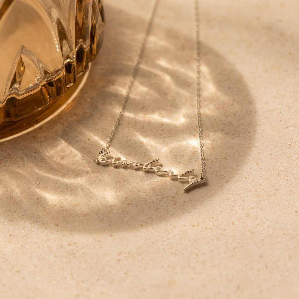 A silver Tiny Venice Name Necklace, custom-made with the name Kimberly, is displayed on a textured surface next to a glass object—a stylish personalized accessory and charming nameplate.