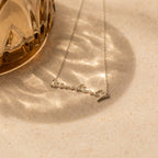 A silver Tiny Venice Name Necklace, custom-made with the name Kimberly, is displayed on a textured surface next to a glass object—a stylish personalized accessory and charming nameplate.