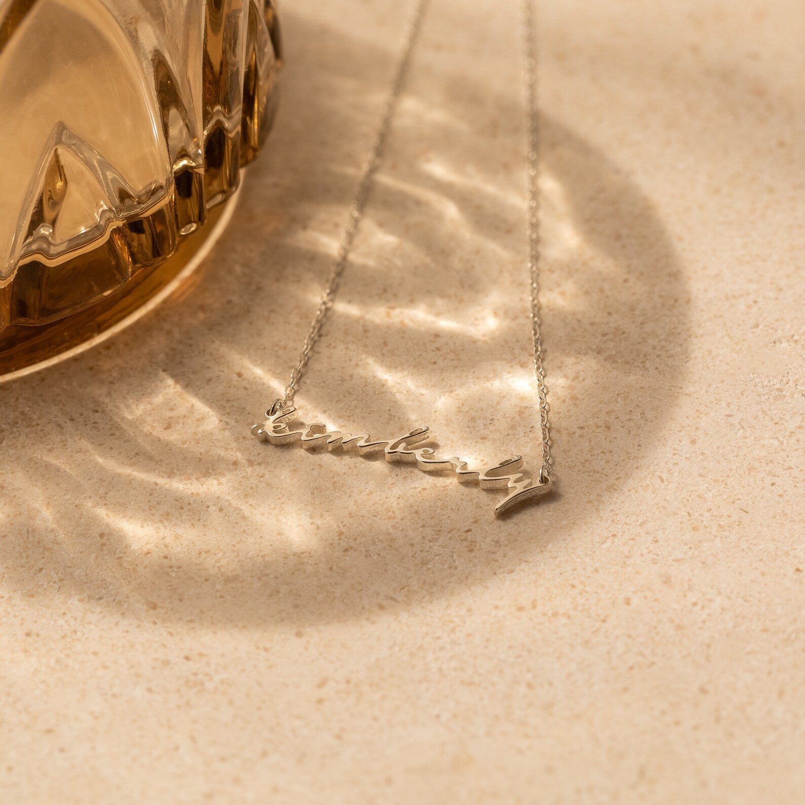A silver Tiny Venice Name Necklace, custom-made with the name Kimberly, is displayed on a textured surface next to a glass object—a stylish personalized accessory and charming nameplate.