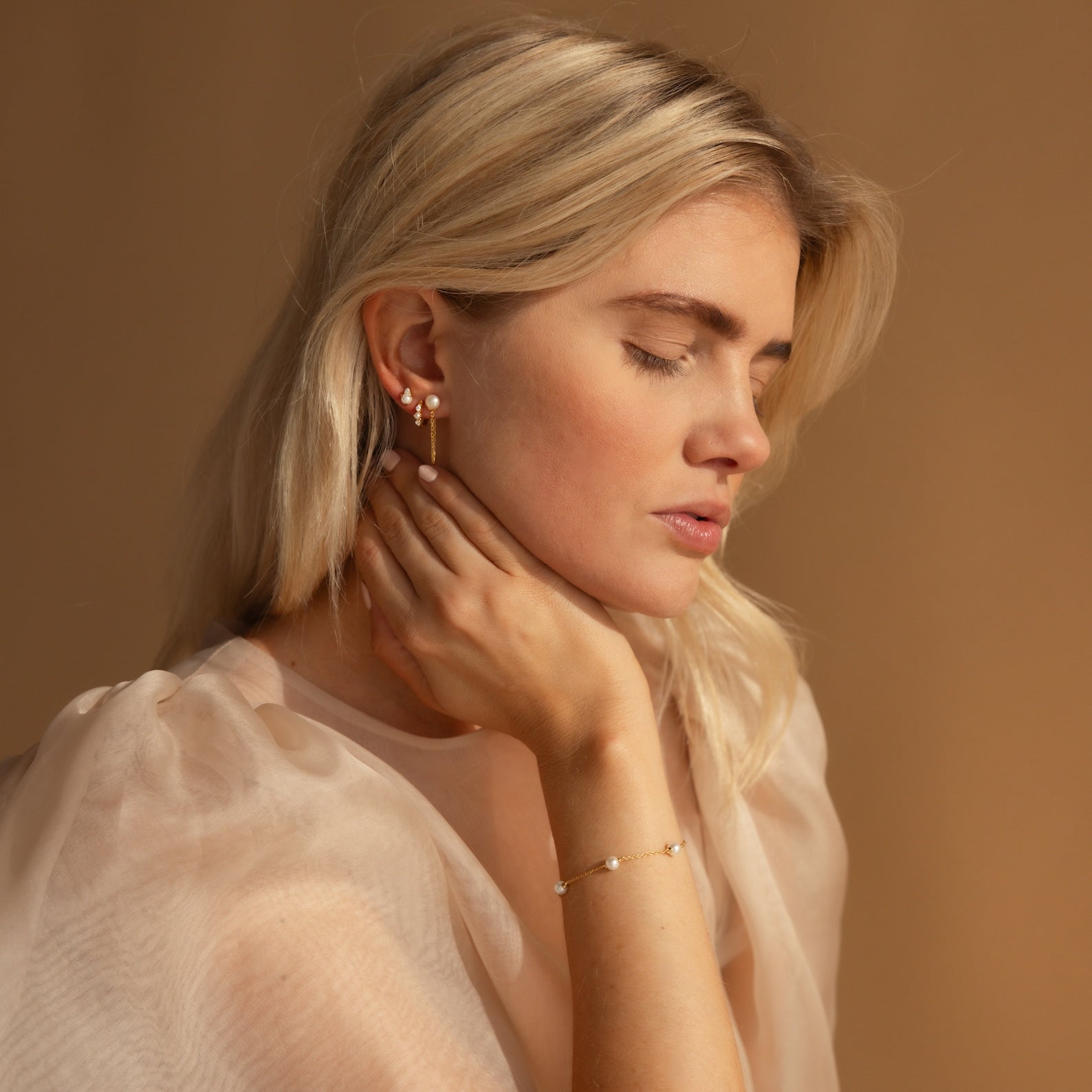 A blonde woman with closed eyes gracefully touches her neck, wearing pearl earrings and the Pearl Station Bracelet in Sterling Silver.