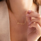 A person gently touches their Tiny Venice Name Necklace, a gold cursive nameplate charm pendant, while wearing it.