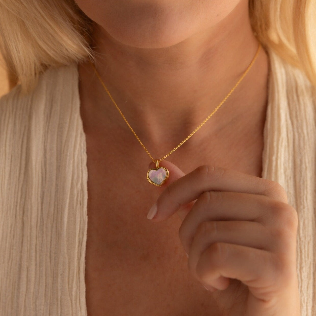 A woman wearing a cream-colored top holds the Pearl Heart Locket Necklace, a gold chain featuring a Mother of Pearl heart locket, near her neck.