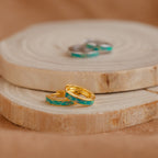 Two Green Opal Inlay Huggies rest on a wooden slice; green earrings and silver styles can be seen blurred in the background.