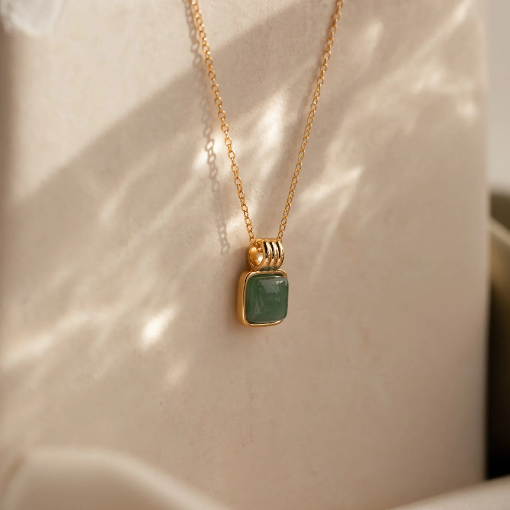 The Evergreen Necklace Set features a gold chain with a square green gemstone pendant, elegantly showcased against a soft beige background with gentle light reflections.