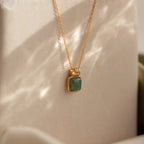 The Evergreen Necklace Set features a gold chain with a square green gemstone pendant, elegantly showcased against a soft beige background with gentle light reflections.