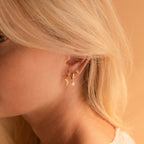 Close-up of a woman’s ear adorned with three gold earrings—a huggie hoop, a moon, and a stud—highlighted by the elegant Pink Opal Drop Huggies, set against a soft peach background.