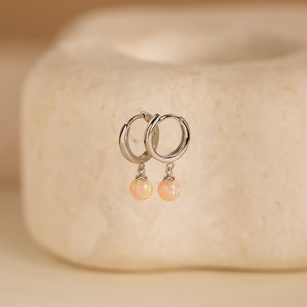 Pink Opal Drop Huggies, featuring silver huggie hoops with dangling pink opal gemstones, are showcased on a smooth beige stone background to add a radiant touch of charm for any occasion.