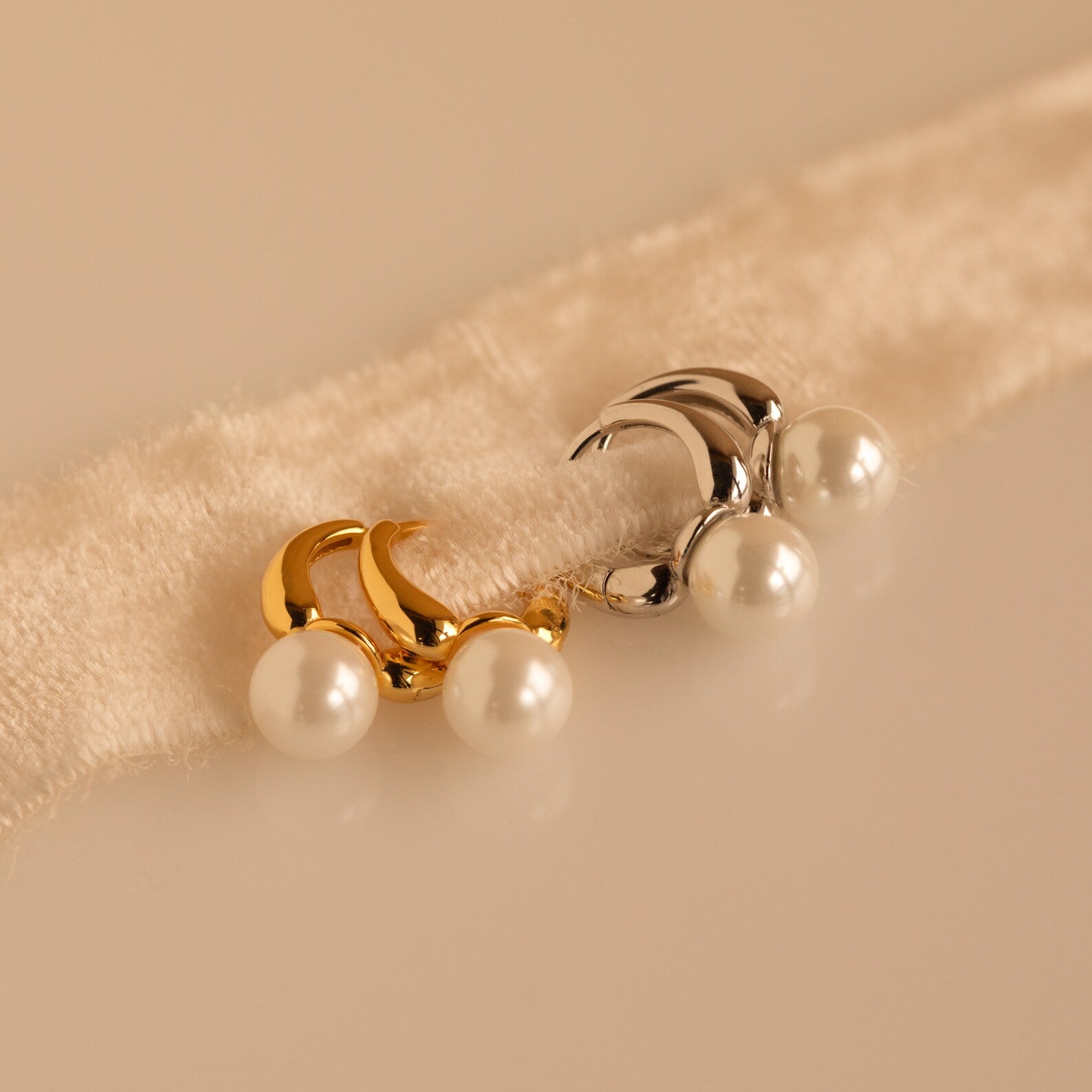 A pair each of gold and silver Pearl Drop Huggies resting on a plush, cream-colored fabric band