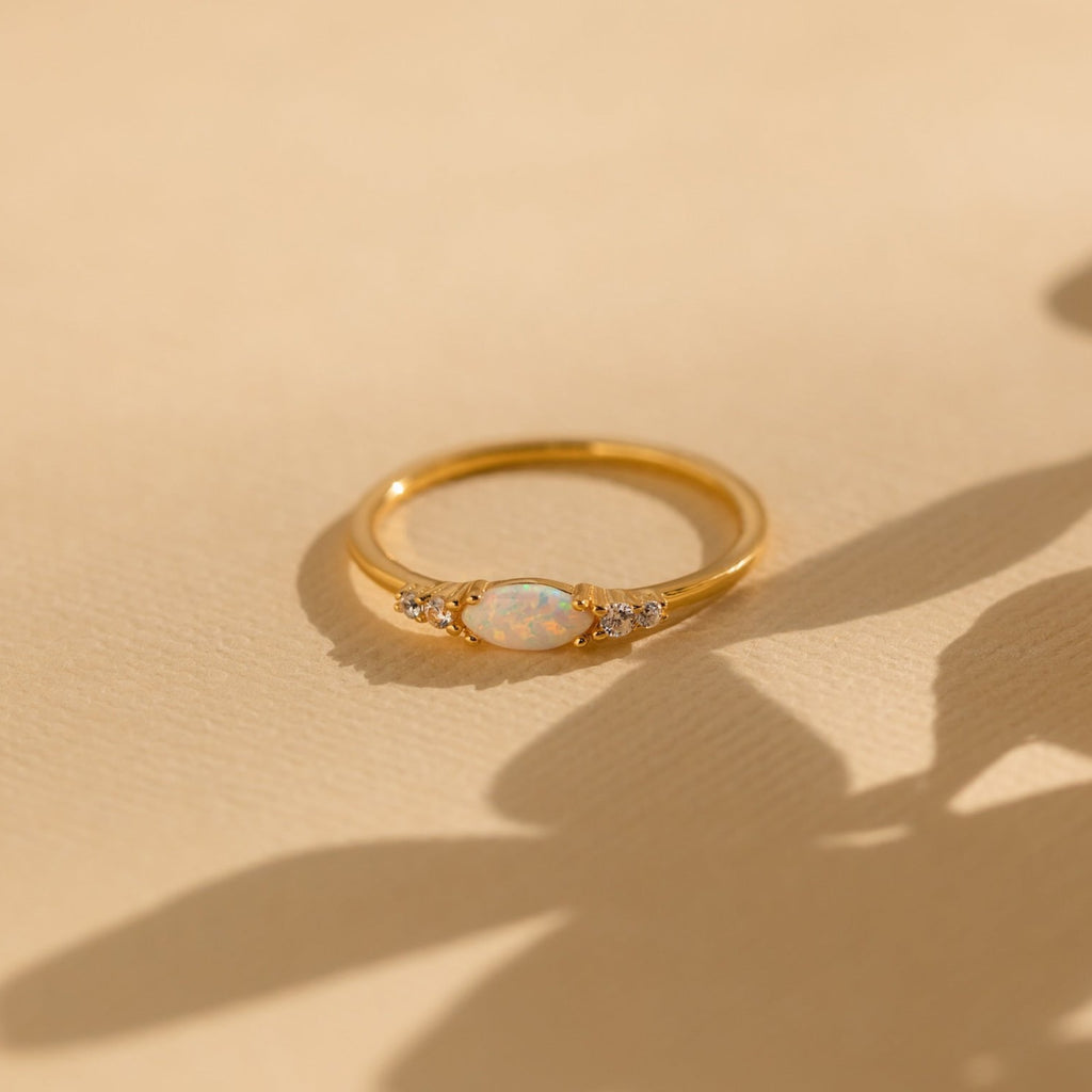 The Marquise Opal Diamond Ring features a gold band with a marquise opal center stone and two small diamonds on each side, displayed on a beige textured surface.
