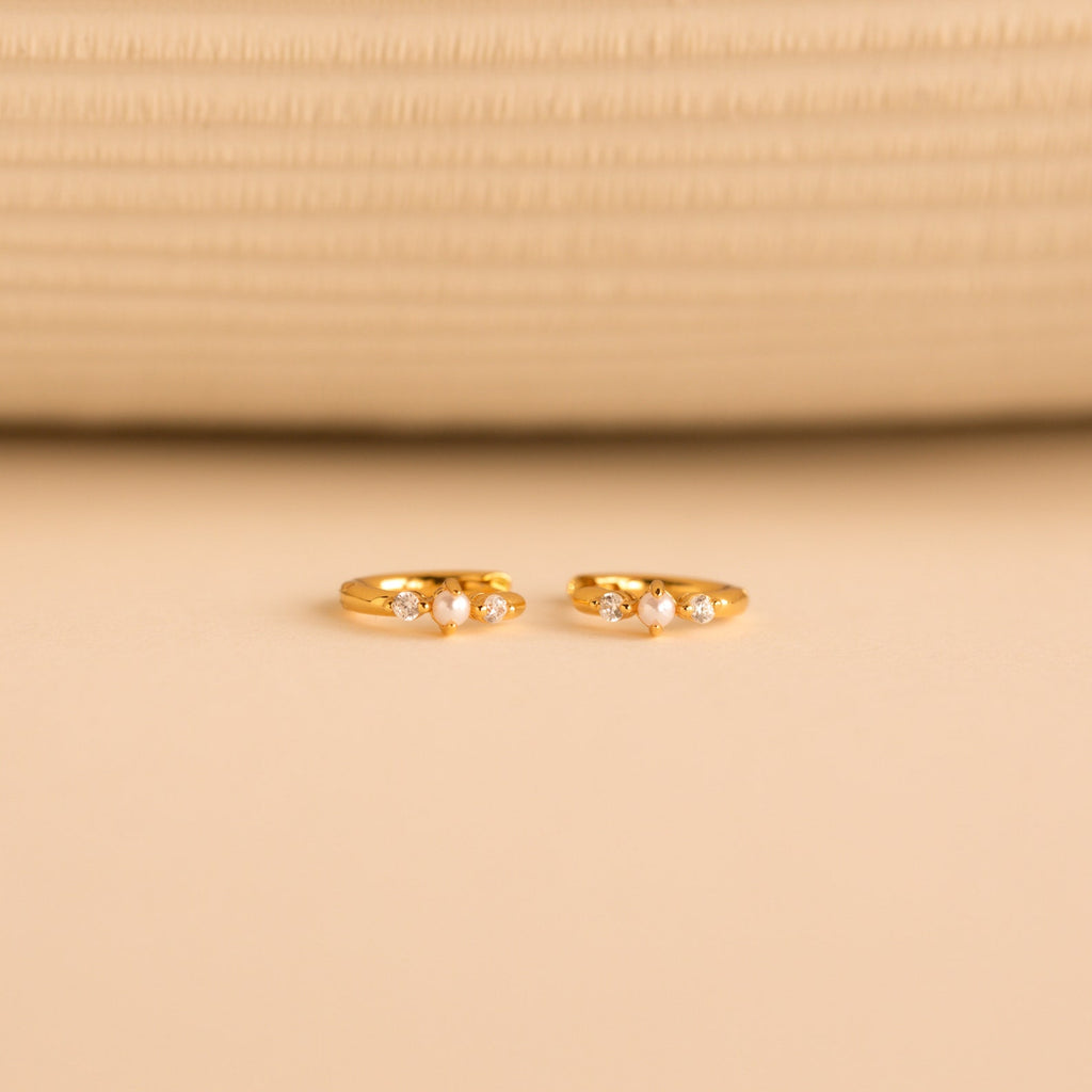 The gold huggies laid flat on a neutral surface, pearls and diamonds prominently featured in the design.