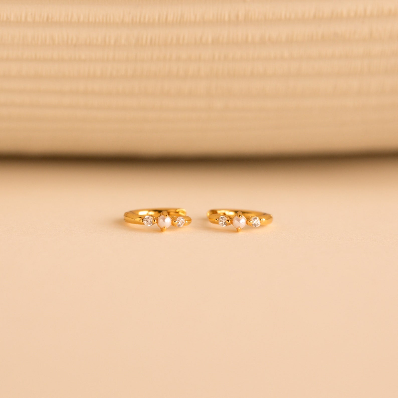 The gold huggies laid flat on a neutral surface, pearls and diamonds prominently featured in the design.