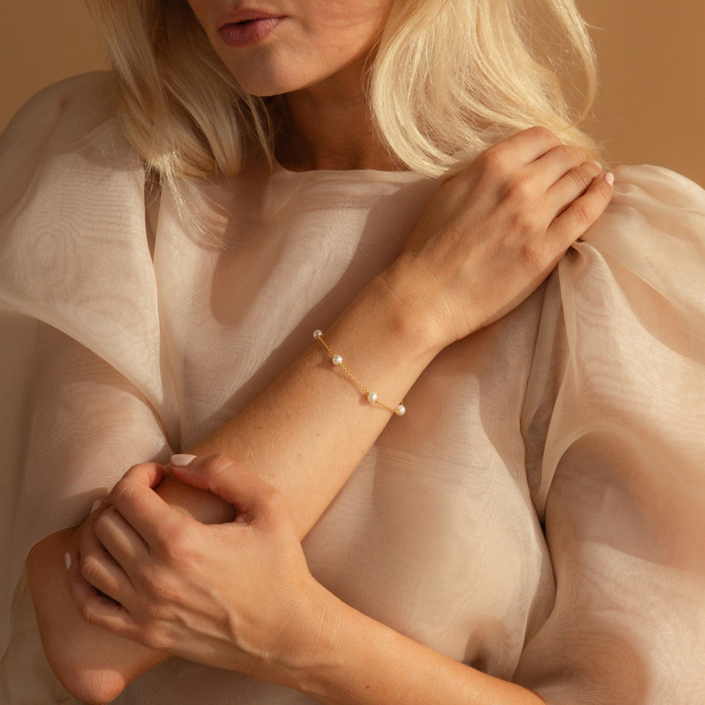 A woman in a sheer beige blouse wears the Pearl Station Bracelet, showcasing modern pearl jewelry as she gently holds her arm.