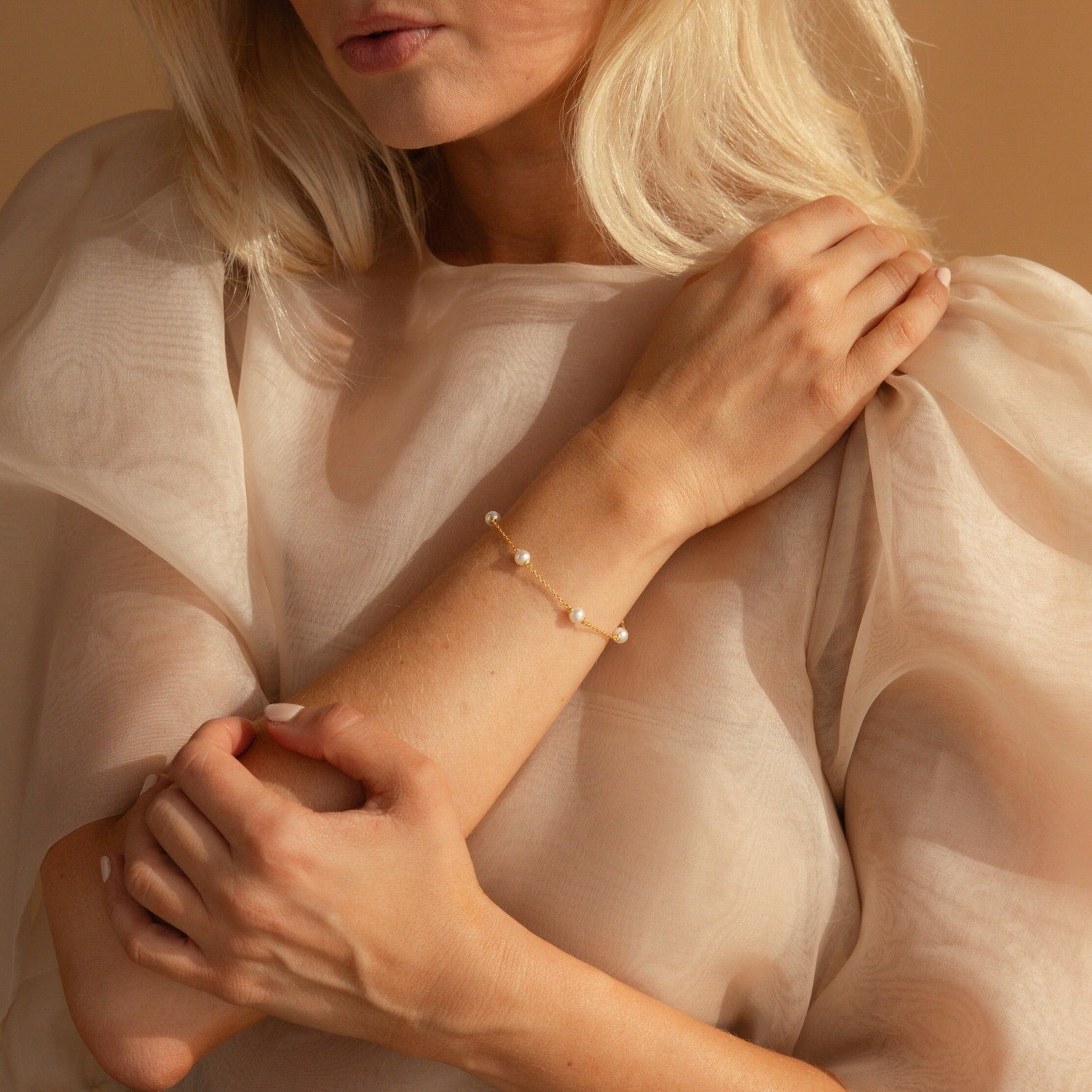 A woman in a sheer beige blouse wears the Pearl Station Bracelet, showcasing modern pearl jewelry as she gently holds her arm.