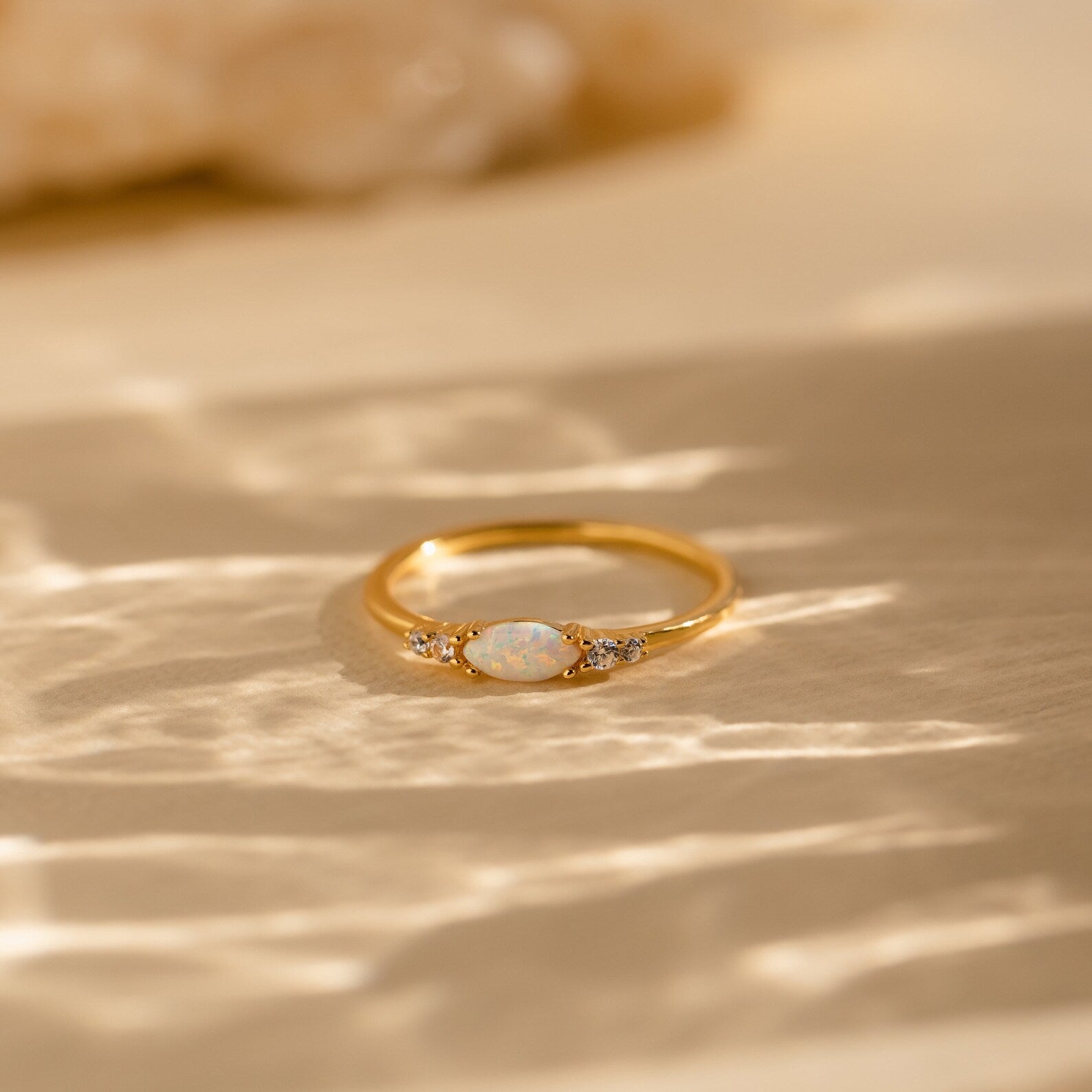 The Marquise Opal Diamond Ring features a central opal with sparkling diamond side stones, beautifully displayed on a textured, sunlit surface.