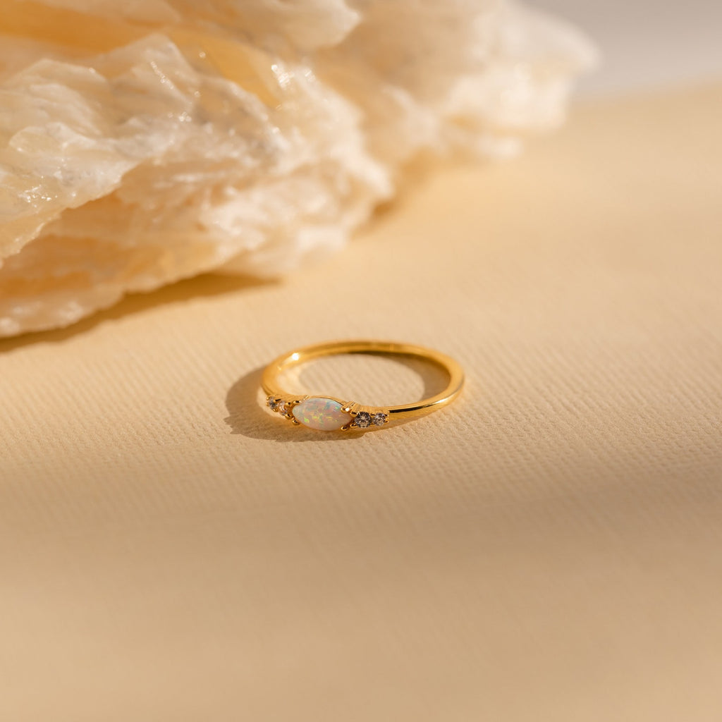 The Marquise Opal Diamond Ring, adorned with small diamonds, rests on a beige surface beside a textured stone.