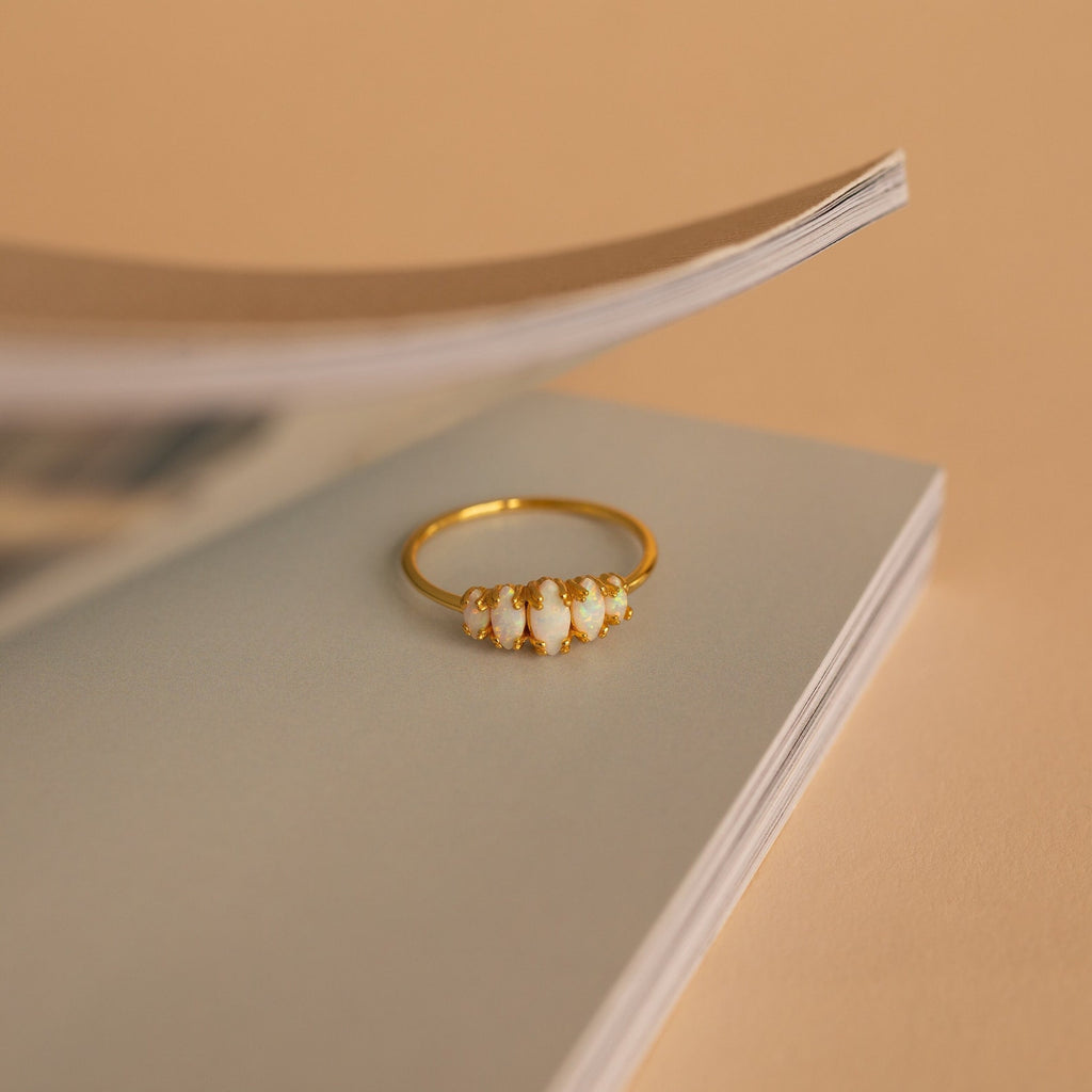 The Marquise Opal Statement Ring with white stones sits on the edge of an open book against a beige background, exuding vintage charm.