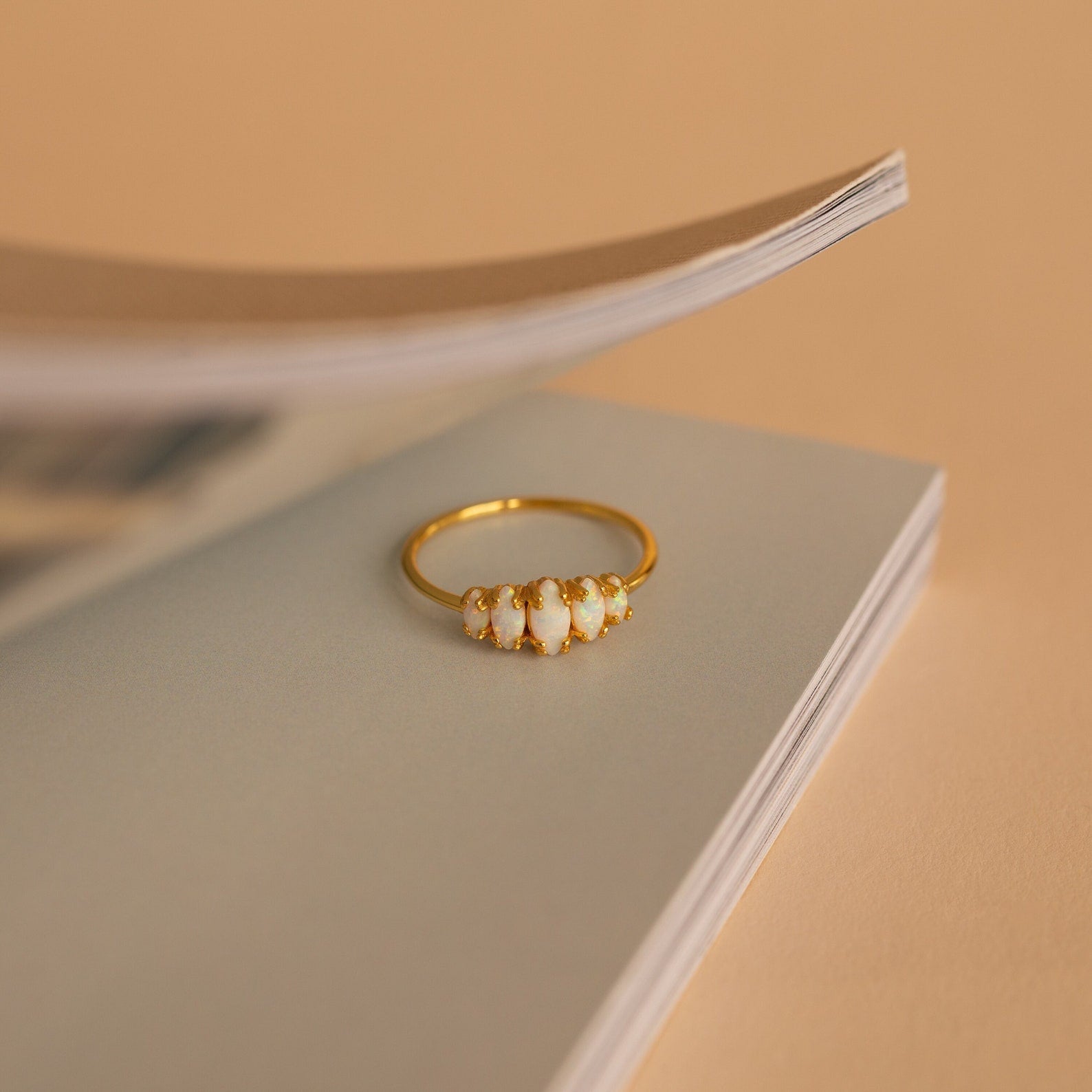 The Marquise Opal Statement Ring with white stones sits on the edge of an open book against a beige background, exuding vintage charm.