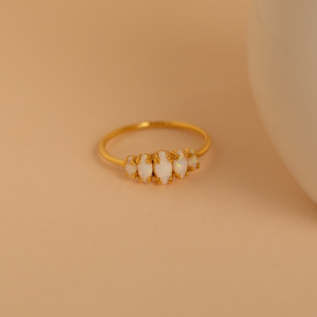 The Marquise Opal Statement Ring features four irregular white stones set in gold, radiating vintage charm, and is displayed on a beige surface next to a round white object.