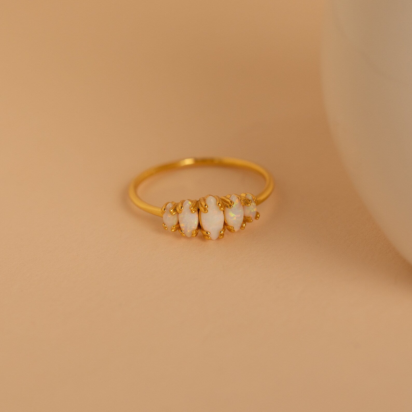 The Marquise Opal Statement Ring features four irregular white stones set in gold, radiating vintage charm, and is displayed on a beige surface next to a round white object.