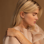 Blonde woman wearing Pearl Chain Studs and a gold bracelet, dressed in a sheer beige top with eyes closed, posing against a tan background.